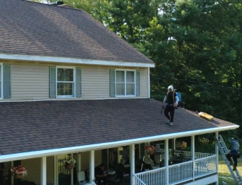 A roofer walking a finished shingle roof above a wrap-around porch on a wooded property